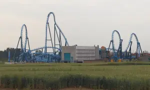 Niebieska kolejka górska Energylandia Abyssus z pętlami i zakrętami na tle pola zboża i budynków.