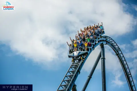 Ludzie krzyczą z radości na pokładzie Hyperiona, największego rollercoastera w Polsce, pędząc po stalowych torach na tle błękitnego nieba.