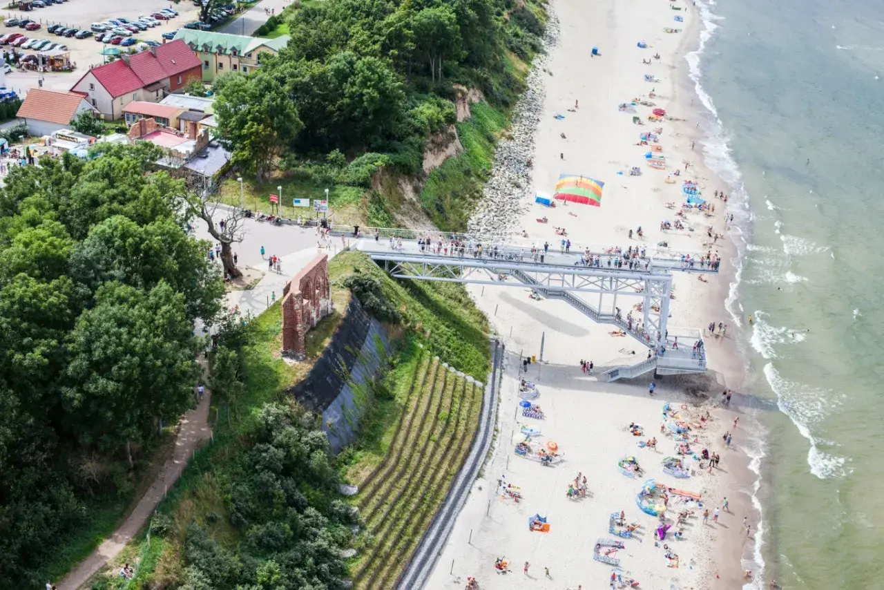 Widok na plażę z kładką i ruinami. Ludzie odpoczywają na piasku, ciesząc się atrakcjami nad morzem.