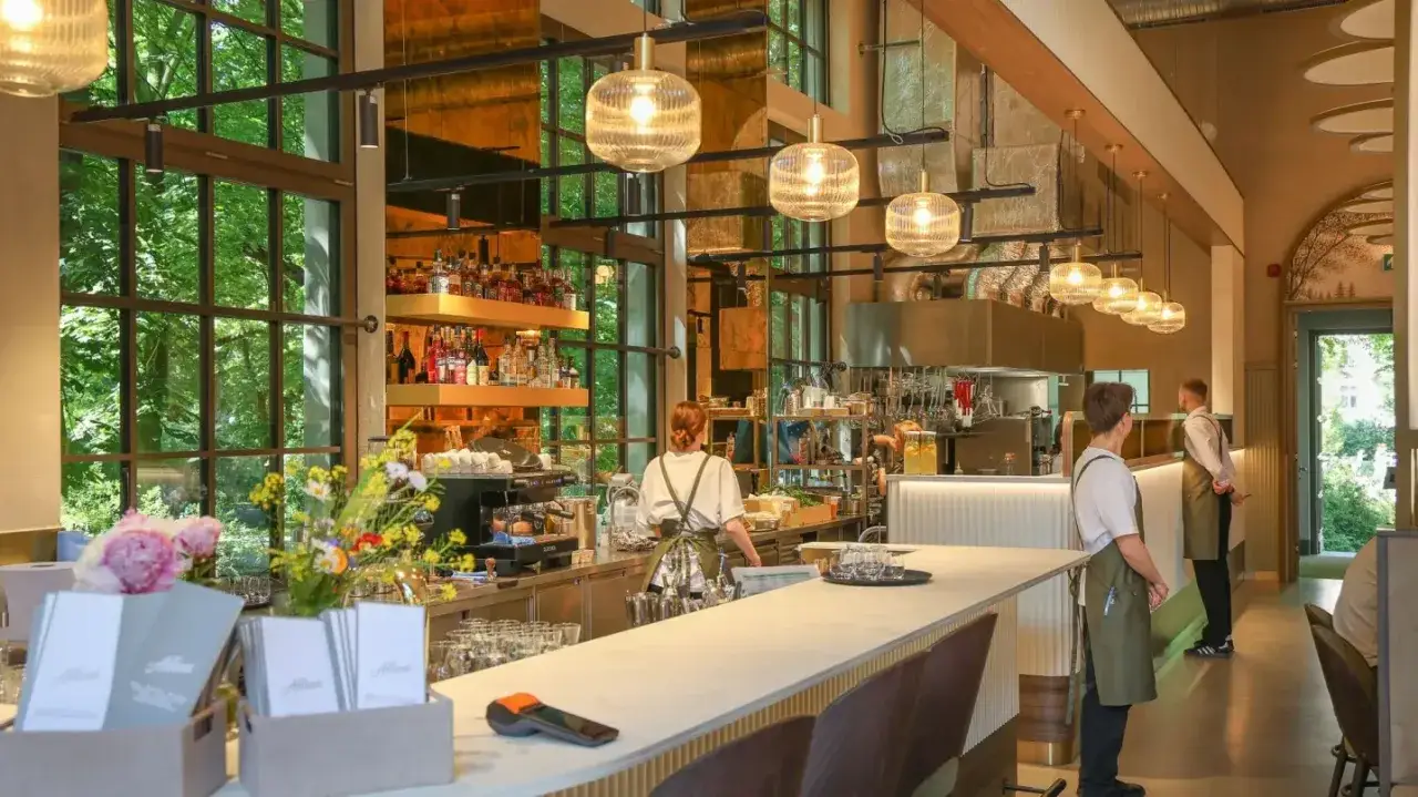 Nowoczesna restauracja z barem, przeszklonymi oknami i zielenią na zewnątrz. Kelnerzy czekają na gości.